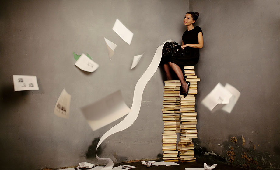 Young female sitting on top of a large pile of books while using a typewriter