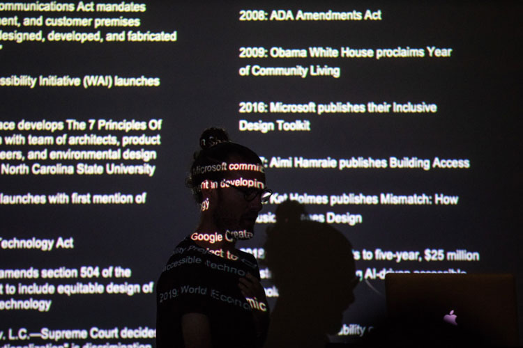 Josh walking in front of a slide which is projected on top of his face. Almost completely black, his figure is silhouetted and a timeline of significant disability design events are featured on the screen including the passage of the ADA and Microsoft's r