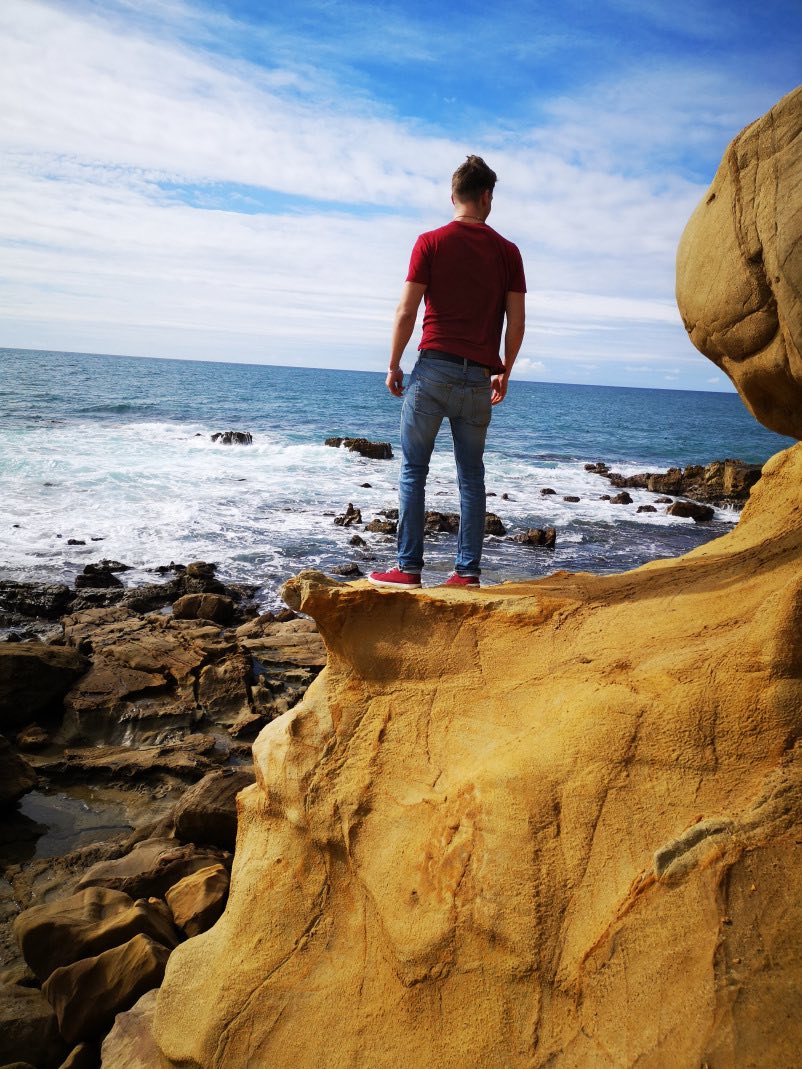 Markus standing at the edge of a cliff, with his back turned to face the rocky shore of an ocean. 