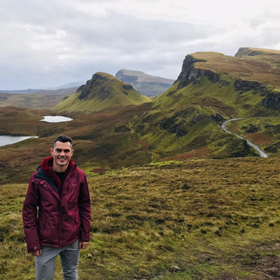 Post-Bacc Counseling graduate Michael Sanchez on a trip with valleys, winding road and mountains in the background in the Quiraing at the Isle of Skye, Scotland