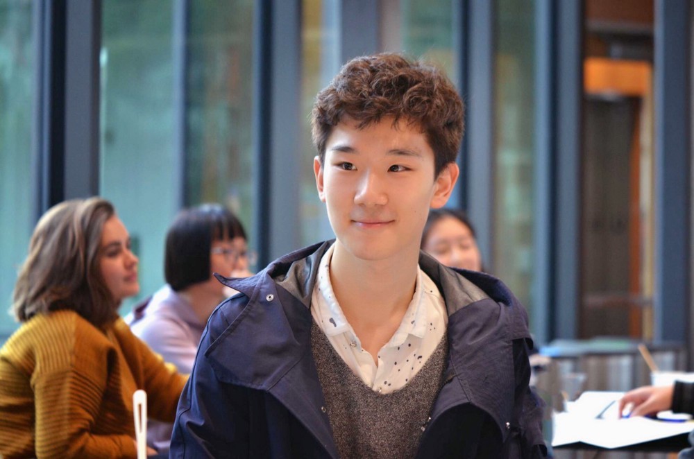 Wenbo at BHGAP orientation, smiling and looking at something beyond the frame, with other students in the background