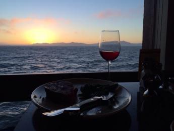 A half-eaten meal paired with a wine glass perched atop a table, photographed with a view of the sun setting on the ocean through a window 