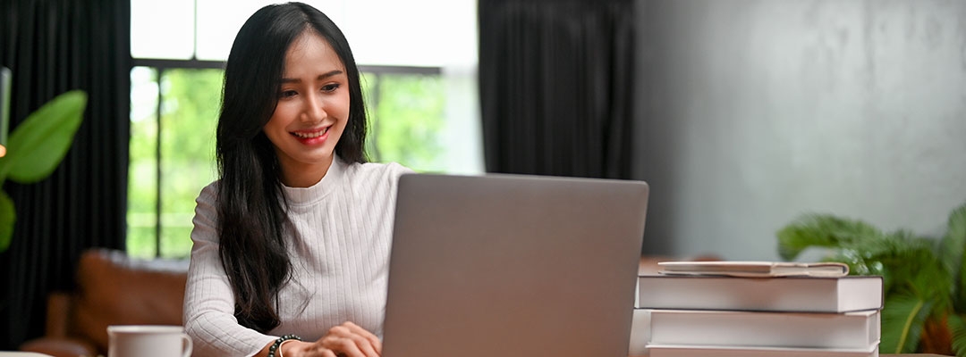 Stock image of a female Asian writer writing on laptop with books on table