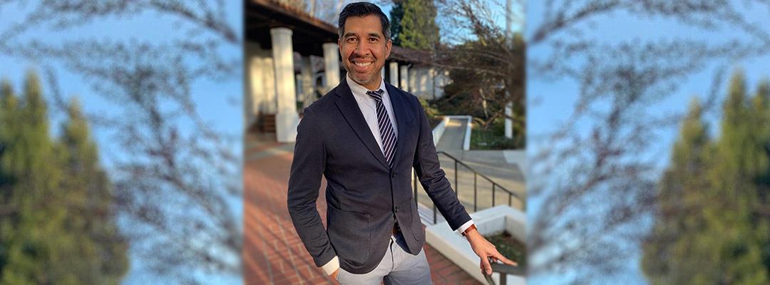 College Admissions and Career Planning certificate program instructor Armando Diaz in blazer and tie, standing next to railing with pillars in background. Photo.