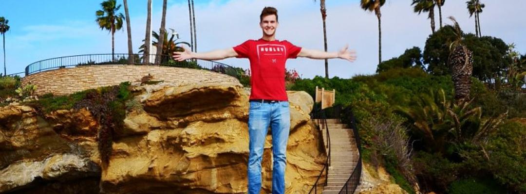 Markus smiling and standing on a rock with his arms outstretched, with palm trees and some greenery in the background of Huntington Beach