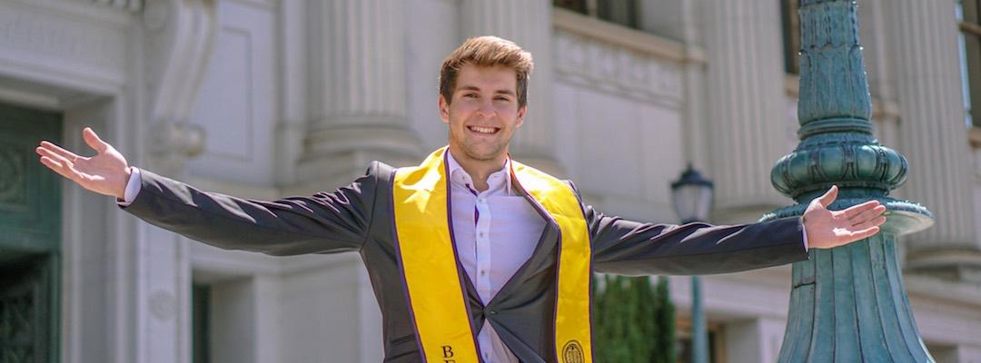Markus sits atop a ledge in front of Doe Library wearing a suit and the Berkeley graduation stole, smiling with his arms outstretched 