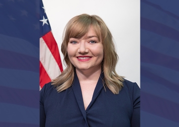 Headshot of Lacy Kelly Ramos in front of U.S. flag on blue background