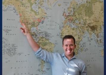 Alex Budak in front of a map of the world, pointing to Berkeley, California