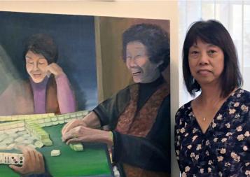 Certificate graduate Elinor Cheung in front of her painting depicting laughing Chinese women enjoying a game of mah-jongg