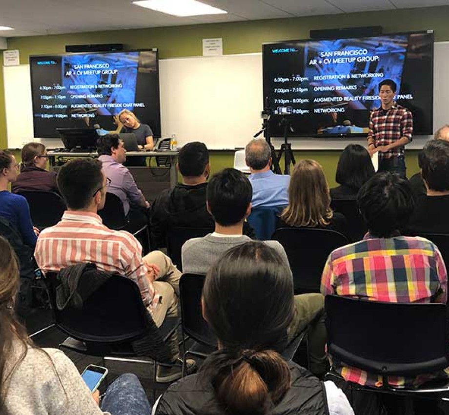 Audience seated and watching a presentation from moderator Sheng Huang in front of a plasma display