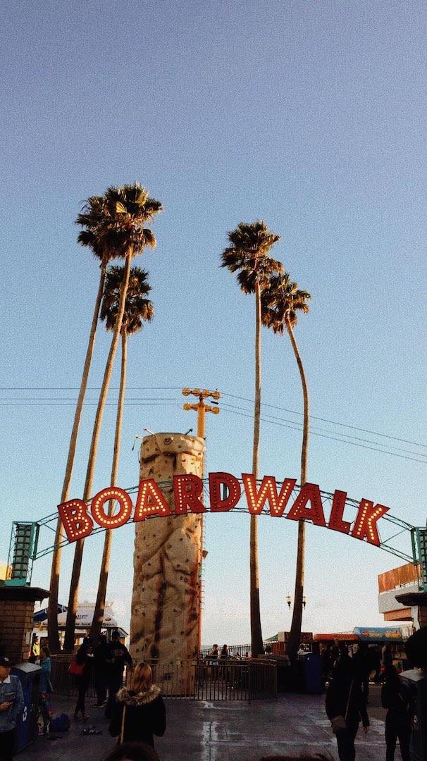 The Santa Cruz Boardwalk entryway sign, taking during the evening and with looming palm trees and people walking around pictured behind
