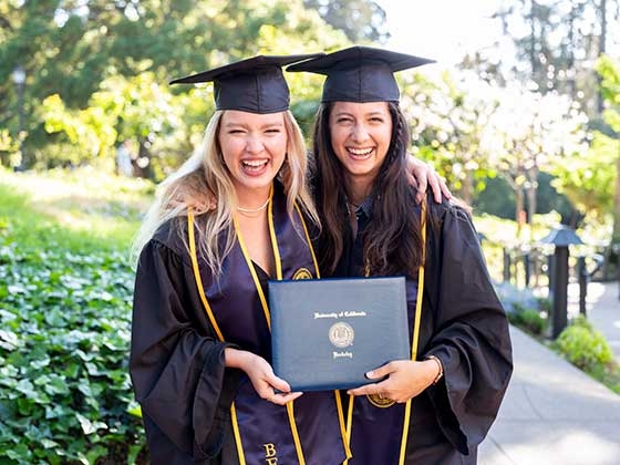 Perline Hochart and friend grin at camera while holding diplomas