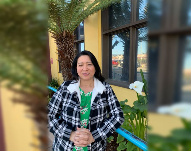 Carmel Gacho in plaid jacket in front of a tree and yellow building