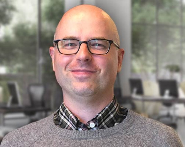 Christopher Hubbard posing while wearing a cardigan over a plad button-down shirt in front of large windows and computer stations  