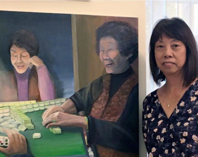 Certificate graduate Elinor Cheung in front of her painting depicting laughing Chinese women enjoying a game of mah-jongg