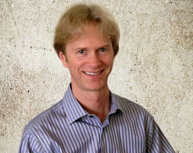 Mark Schrier posing in front of a concrete wall wearing a striped button down shirt