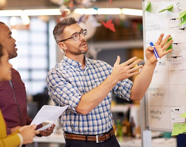 Man pointing at whiteboard with sticky notes while 2 colleagues look on