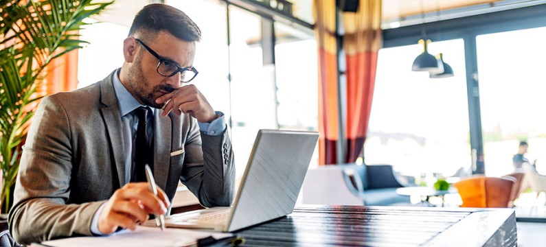 Serious millennial man using laptop sitting at cafe table, focused guy in suit communicating online, writing emails, distantly working or studying on computer in public place.